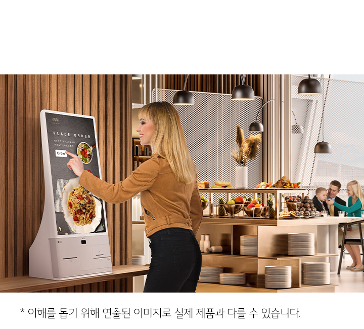 한 여성이 터치스크린 키오스크를 사용하고 있습니다. 화면에는 PLACE ORDER, BEST ITALIAN RESTAURANT, Order라는 텍스트가 보이며, 이탈리안 요리가 이미지로 표시되어 있습니다. 배경에는 식당 내부가 보이며, 테이블 위에 다양한 과일과 빵이 놓여 있습니다. 가족으로 보이는 사람들이 테이블에 앉아 대화를 나누고 있습니다. 하단에는 이해를 돕기 위해 연출된 이미지로 실제 제품과 다를 수 있습니다.라는 문구가 있습니다.
