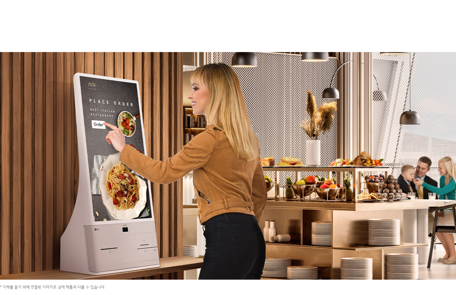 한 여성이 터치스크린 키오스크를 사용하고 있습니다. 화면에는 PLACE ORDER, BEST ITALIAN RESTAURANT, Order라는 텍스트가 보이며, 이탈리안 요리가 이미지로 표시되어 있습니다. 배경에는 식당 내부가 보이며, 테이블 위에 다양한 과일과 빵이 놓여 있습니다. 가족으로 보이는 사람들이 테이블에 앉아 대화를 나누고 있습니다. 하단에는 이해를 돕기 위해 연출된 이미지로 실제 제품과 다를 수 있습니다.라는 문구가 있습니다.