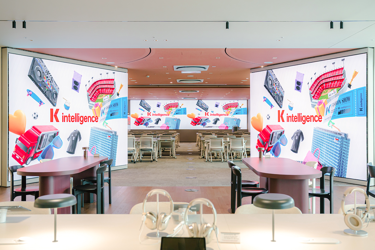 대형 사이니지가 정면과 좌우 벽면에 설치된 행사 공간에서 여러 줄로 배치된 회의용 의자와 테이블이 중앙에 보이고, 모든 사이니지에 붉은색 글자 'K intelligence'가 크게 표시되며 주변에 스피커, 경기장, 트럭, 건물, 의류, 티켓 등의 그래픽이 배치되어 있고, 전경에는 헤드폰과 태블릿이 놓인 체험 테이블이 보입니다.