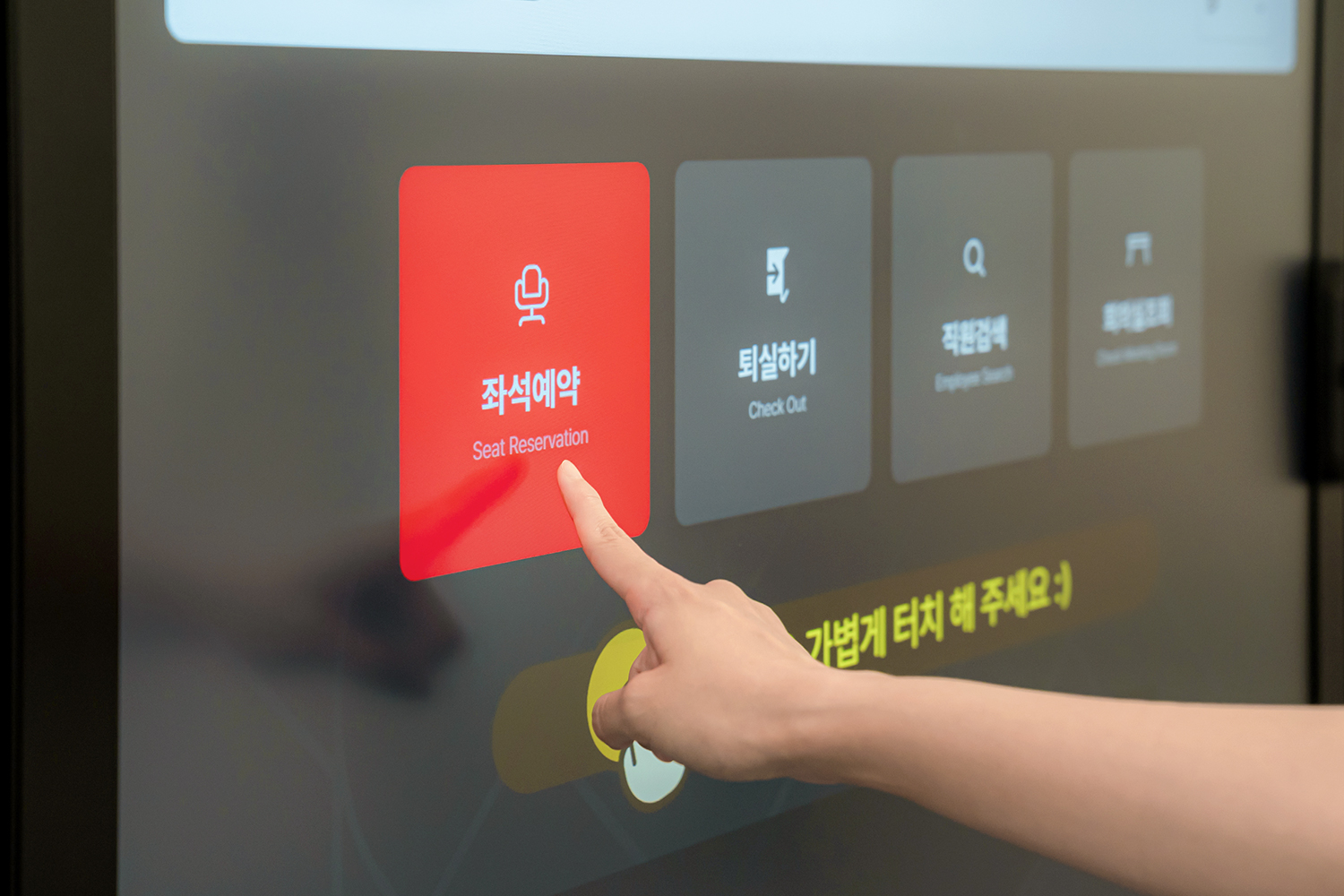 손가락이 사이니지의 터치스크린에 있는 붉은색 버튼을 누르는 장면으로, 버튼에는 한글 '좌석예약'과 영문 'Seat Reservation'이 함께 쓰여 있고, 오른쪽 인접 버튼들에는 '퇴실하기'와 'Check Out', '직원검색'과 'Employee Search'가 각각 표시되어 있습니다.