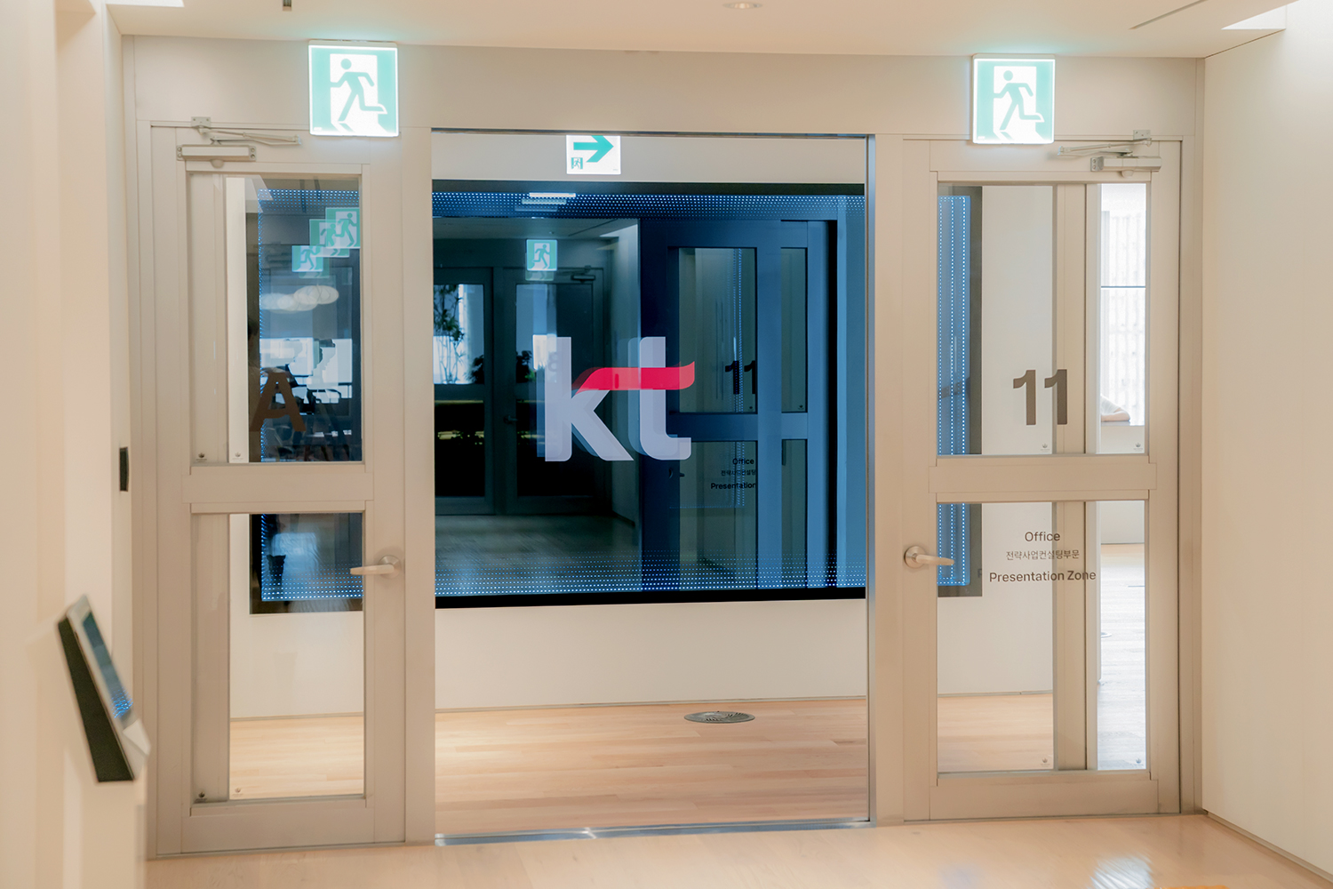 숫자 '11'이 적힌 유리 출입문 너머로 대형 사이니지에 'KT' 로고가 중앙에 보이고, 오른쪽 유리문에는 'Office'와 'Presentation Zone' 문구가 인쇄되어 있으며, 문 상단에는 비상구 표지판이 설치되어 있습니다.