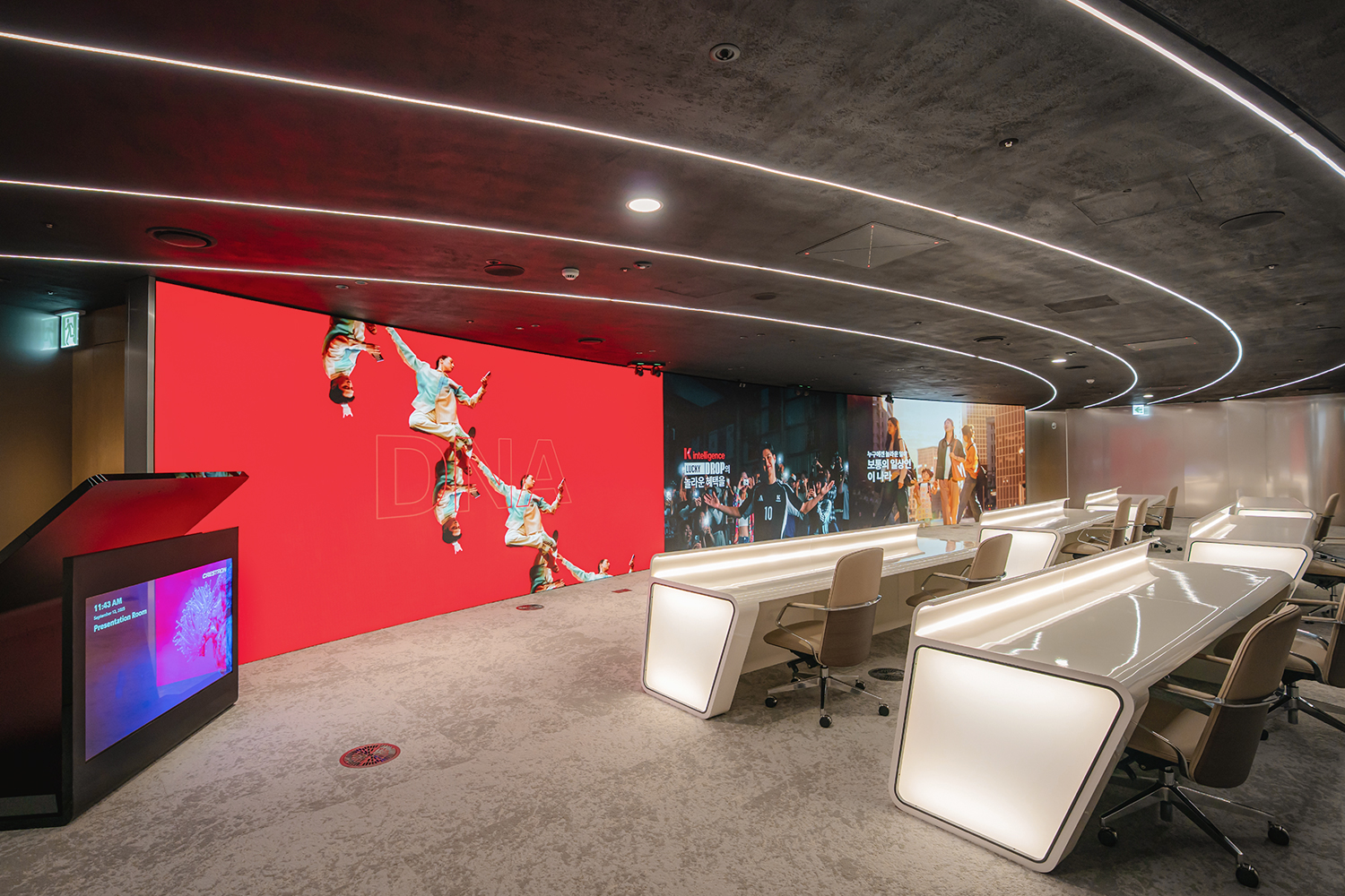 천장에 곡선 라인 조명이 있는 프레젠테이션 룸에서 대형 사이니지 화면에 붉은 배경과 반투명 문자 'DNA'가 있고 인물 사진과 설명 텍스트가 이어져 있으며, 앞쪽에 바퀴 달린 회의용 의자와 테이블이 배치되어 있습니다.