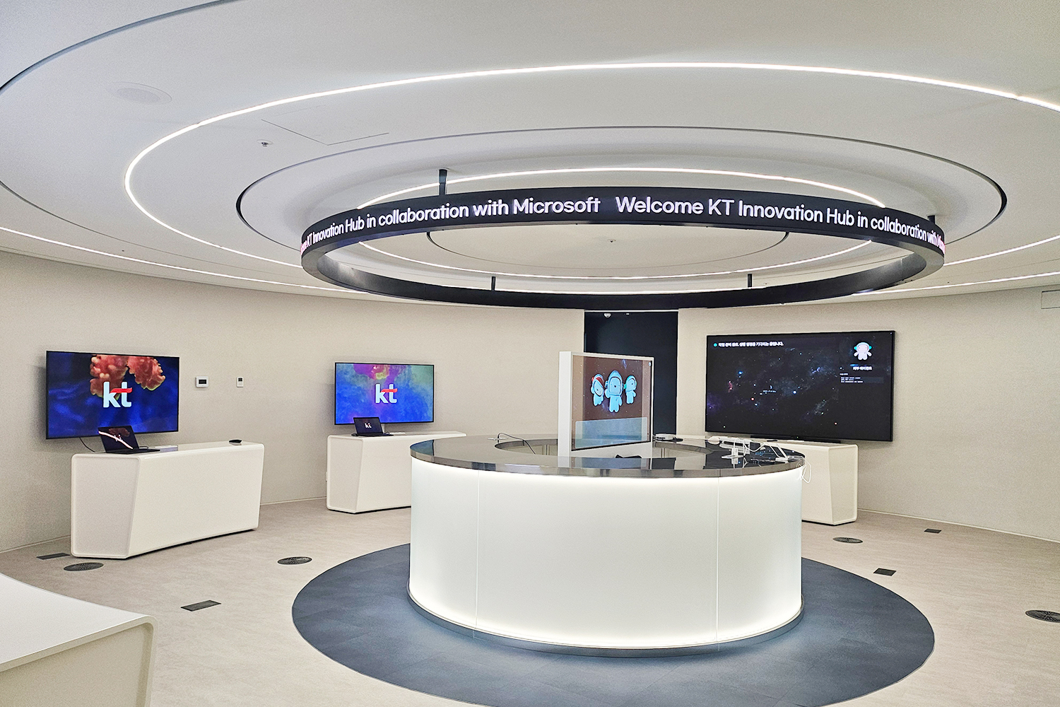 원형 천장 구조물에 둘러진 띠형 디스플레이에 'Welcome KT Innovation Hub in collaboration with Microsoft'가 반복 표기되고, 실내 벽면 모니터에는 'KT' 로고 화면이 보이며 중앙에는 둥근 안내 데스크와 주변에 전시용 스크린과 노트북이 배치되어 있습니다.