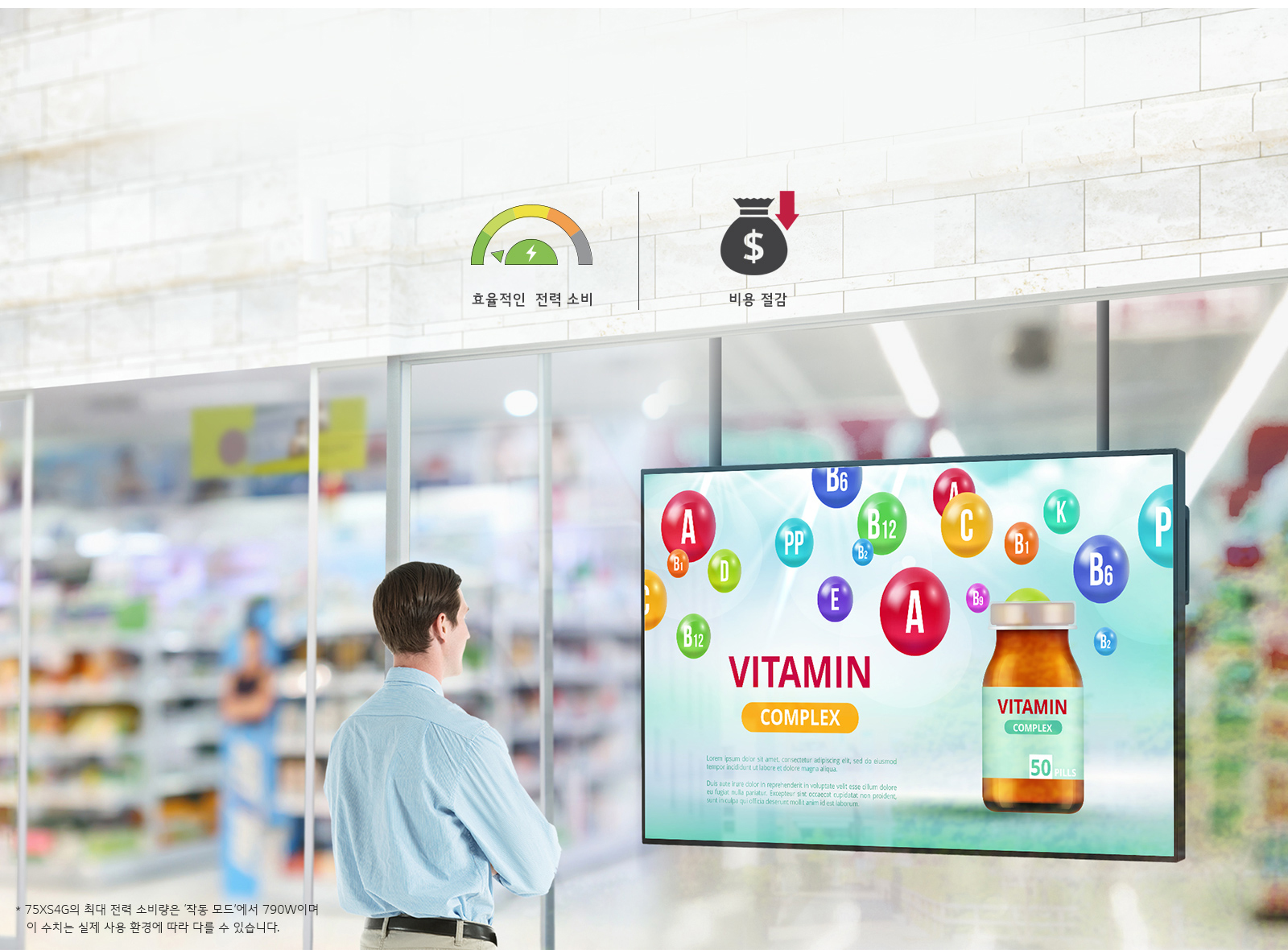 한 남성이 대형 스크린을 보고 있으며, 스크린에는 다양한 비타민 알파벳과 함께 VITAMIN COMPLEX라는 문구와 비타민 병 이미지가 보입니다. 상단에는 효율적인 전력 소비와 비용 절감이라는 문구가 각각 전력 소비 게이지와 비용 절감 아이콘과 함께 있습니다. 하단에는 75XS4G의 최대 전력 소비량은 '작동 모드'에서 790W이며 이 수치는 실제 사용 환경에 따라 다를 수 있습니다.라는 설명이 있습니다. 배경은 상점 내부로 보이며 다양한 상품들이 진열되어 있습니다.