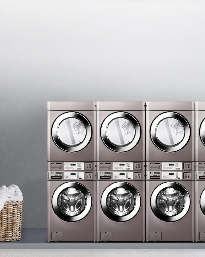세탁기와 건조기가 나란히 배치되어 있는 이미지입니다. 각 기기는 금속성 외관을 가지고 있으며, 디지털 디스플레이와 버튼이 장착되어 있습니다. 세탁기와 건조기 위에는 각각의 문이 투명하게 되어 있어 내부가 보입니다. 이미지의 한쪽에는 빨래 바구니가 놓여 있습니다. 배경은 회색 벽으로 되어 있으며, 한쪽에는 커튼이 드리워져 있습니다.