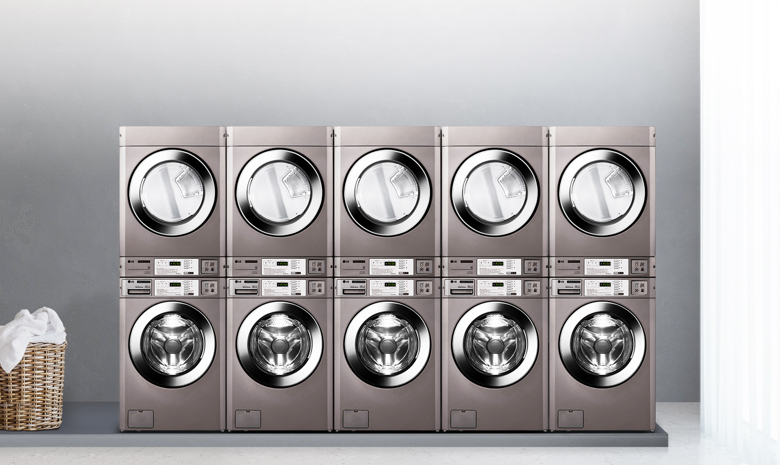 세탁기와 건조기가 나란히 배치되어 있는 이미지입니다. 각 기기는 금속성 외관을 가지고 있으며, 디지털 디스플레이와 버튼이 장착되어 있습니다. 세탁기와 건조기 위에는 각각의 문이 투명하게 되어 있어 내부가 보입니다. 이미지의 한쪽에는 빨래 바구니가 놓여 있습니다. 배경은 회색 벽으로 되어 있으며, 한쪽에는 커튼이 드리워져 있습니다.