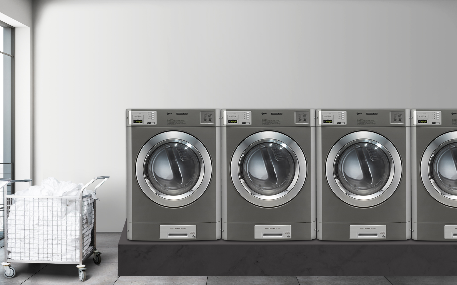 세탁실의 모습입니다. 네 대의 산업용 세탁기가 나란히 놓여 있으며, 각 세탁기의 디스플레이에는 IN USE라는 문구가 표시되어 있습니다. 세탁기 옆에는 흰색 세탁물이 담긴 금속 바구니가 있습니다. 바닥은 타일로 되어 있고, 벽은 밝은 색상입니다. 창문을 통해 자연광이 들어오고 있습니다.