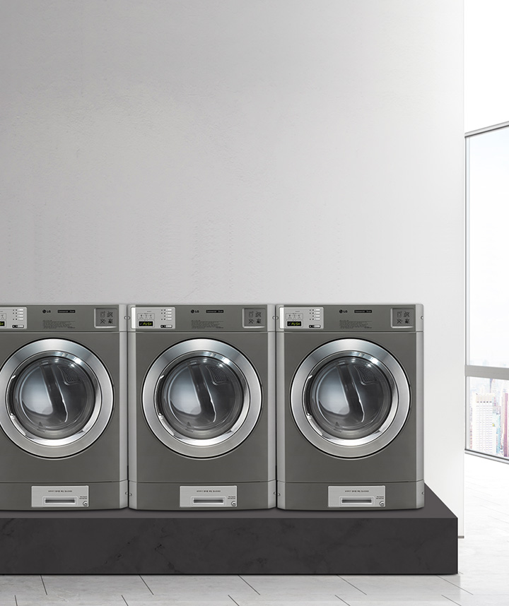 회색 벽 앞에 다섯 대의 산업용 세탁기가 나란히 놓여 있습니다. 세탁기들은 금속성의 외관을 가지고 있으며, 각각의 문은 투명한 원형 창으로 되어 있습니다. 세탁기 상단에는 조작 패널이 있으며, 디지털 디스플레이와 버튼들이 보입니다. 바닥은 대리석 타일로 되어 있습니다. 창문을 통해 도시 풍경이 보입니다.