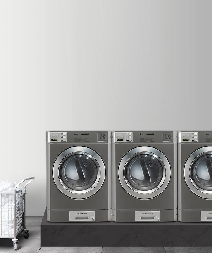 세탁실의 모습입니다. 네 대의 산업용 세탁기가 나란히 놓여 있으며, 각 세탁기의 디스플레이에는 IN USE라는 문구가 표시되어 있습니다. 세탁기 옆에는 흰색 세탁물이 담긴 금속 바구니가 있습니다. 바닥은 타일로 되어 있고, 벽은 밝은 색상입니다. 창문을 통해 자연광이 들어오고 있습니다.