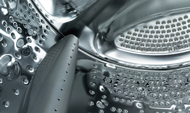 세탁기 드럼 내부의 금속 표면이 보입니다. 표면에는 작은 구멍들이 규칙적으로 배열되어 있으며, 금속의 반짝이는 질감이 강조되어 있습니다. 드럼의 일부에는 물살을 일으키는 돌출된 구조물이 있습니다.