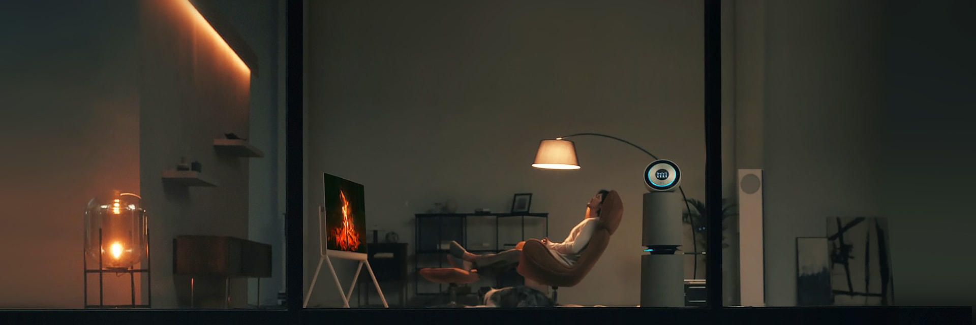 어두운 거실에서 한 여성이 안락의자에 앉아 불꽃 영상이 나오는 TV를 바라보며 휴식을 취하는 모습