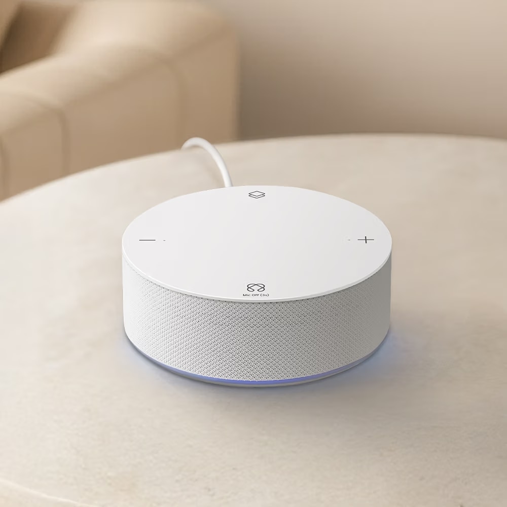 
베이지색 원형 테이블 위에 놓인 심플한 화이트 색상의 원통형 스마트 허브 기기 클로즈업. 기기 상단 평평한 면에는 터치 인터페이스 아이콘이 배치되어 있으며, 12시 방향에는 마름모 꼴의 레이어 아이콘, 9시 방향에는 마이너스(-), 3시 방향에는 플러스(+) 아이콘, 6시 방향에는 무한대(∞) 모양의 아이콘과 그 아래 'Mic Off (3s)'라는 회색 텍스트가 적혀 있음. 기기 측면은 화이트 패브릭 매쉬 소재로 마감되어 있고, 기기 하단 테두리에서는 은은한 파란색 LED 불빛이 들어오고 있음. 배경에는 베이지색 소파가 흐릿하게 보임.