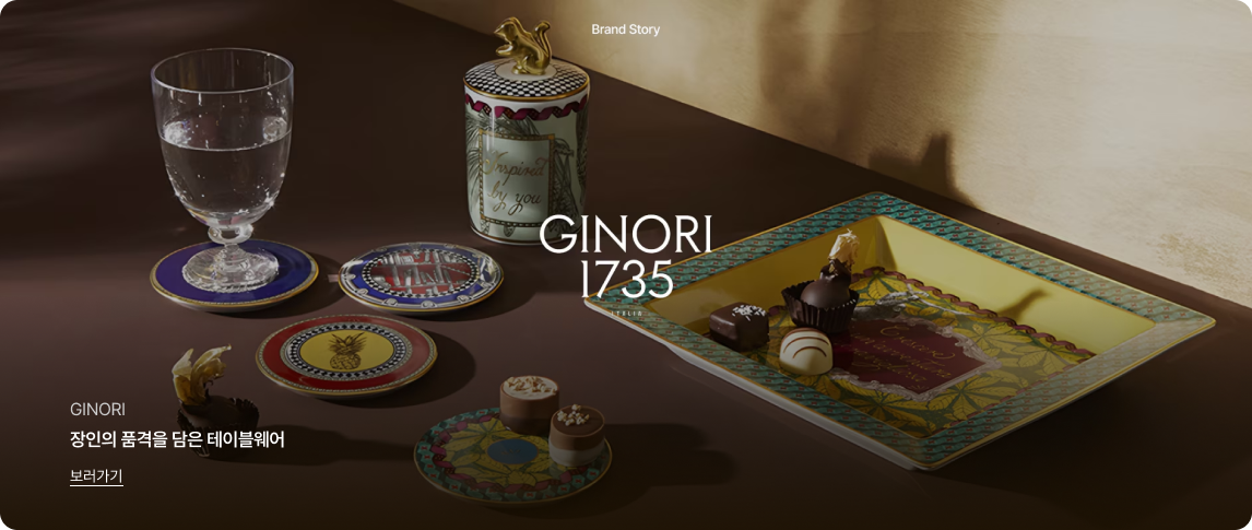 어두운 배경 위로 따뜻한 조명을 받아 우아하게 빛나는 디저트 테이블 세팅 이미지입니다.
이미지 상단 중앙에는 'Brand Story' 문구가 있고, 중앙에는 흰색 'GINORI 1735' 로고가 있습니다.
우측에는 노란색 테두리의 사각 접시 위에 초콜릿이 담겨 있고, 좌측에는 화려한 패턴의 원형 접시들과 'Inspired by you'라고 적힌 뚜껑 있는 도자기, 물이 담긴 투명한 유리잔이 놓여 있습니다.
좌측 하단에는 'GINORI', '장인의 품격을 담은 테이블웨어'라는 문구와 밑줄이 그어진 '보러가기' 링크가 있습니다.