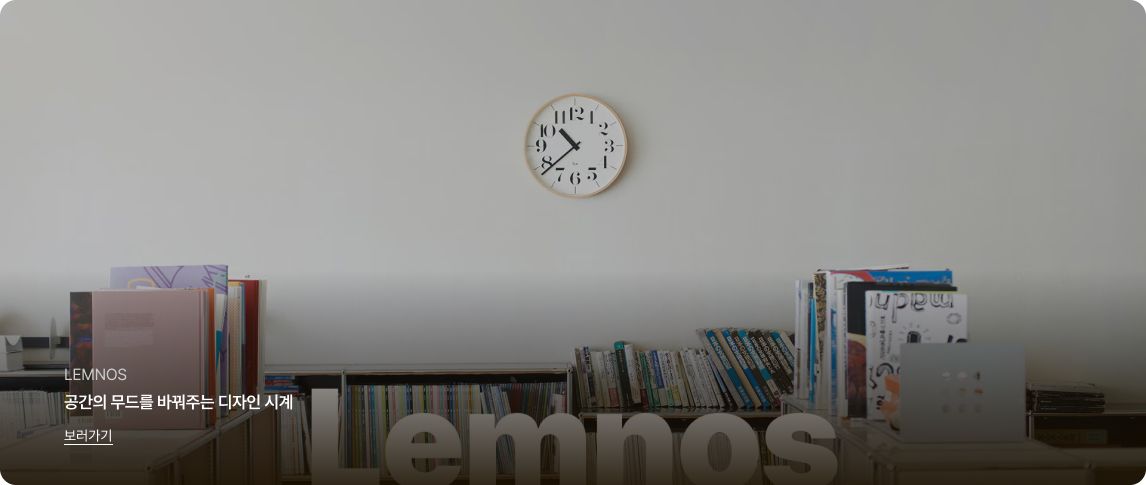 깨끗한 흰색 벽면 중앙에 심플한 디자인의 원형 목재 벽시계가 걸려 있는 미니멀한 공간 이미지입니다.
벽면 아래쪽에는 낮은 책장에 다양한 색상의 책들이 빼곡하게 꽂혀 있습니다. 이미지 하단부에는 큰 영문 타이포그래피로 'Lemnos'라는 글자가 배경처럼 희미하게 깔려 있습니다.
좌측 하단에는 흰색 텍스트로 'LEMNOS', '공간의 무드를 바꿔주는 디자인 시계'라는 문구와 밑줄이 그어진 '보러가기' 링크가 있습니다.