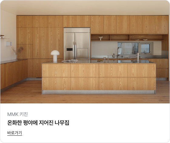 공간 전체를 아우르는 밝은 우드 톤의 수납장이 벽면과 아일랜드 식탁에 적용되어 따뜻하고 아늑한 느낌을 주는 주방 이미지입니다. 수납장 중앙에는 스테인리스 재질의 냉장고가 빌트인 되어 있고, 싱크대 상판 위에는 작은 버섯 모양의 조명이 놓여 있습니다.
하단에는 'MMK 키친', '온화한 평야에 지어진 나무집'이라는 제목과 '바로가기' 링크가 있습니다.
