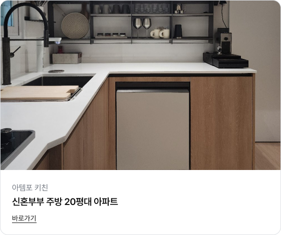 따뜻한 우드 톤의 하부장과 깨끗한 화이트 상판으로 꾸며진 아늑한 주방의 코너 공간 이미지입니다. 하부장에는 베이지색 식기세척기가 빌트인 되어 있고, 벽면에는 블랙 철제 프레임의 선반이 설치되어 와인잔과 컵들이 가지런히 정리되어 있습니다. 검은색 수전이 포인트가 되어 세련미를 더합니다.
하단에는 '아템포 키친', '신혼부부 주방 20평대 아파트'라는 제목과 '바로가기' 링크가 있습니다.