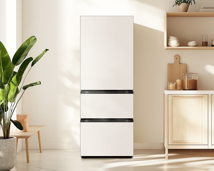 햇살이 들어오는 밝은 실내 중앙에 상칸은 도어형, 중·하칸은 서랍형 구조인 흰색 김치냉장고가 배치되어 있고 좌측에는 큰 화분, 우측에는 우드 톤의 수납장이 있는 이미지