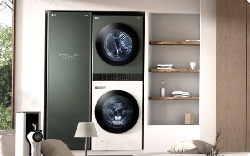 좌측에는 짙은 녹색(카밍 그린)의 LG 스타일러가 빌트인 되어 있고, 그 옆으로 상단 건조기는 짙은 녹색, 하단 세탁기는 화이트 색상으로 구성된 일체형 LG 워시타워가 나란히 배치되어 있음. 워시타워 우측에는 3단 우드 선반이 있는 벽면 수납공간에 흰색 수건과 소품들이 정돈되어 있으며, 하단 좌측에는 LG 코드제로 무선 청소기의 일부와 현대적인 디자인의 테이블 스탠드가 놓여 있는 세탁실 인테리어 사진.