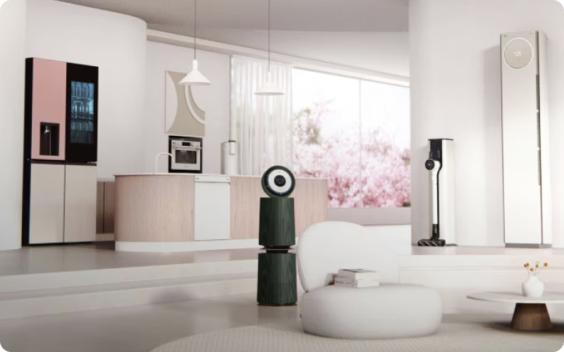 밝고 화사한 햇살이 들어오는 거실 인테리어 사진. 좌측에는 핑크색 패널과 내부가 보이는 노크온 도어가 적용된 LG 오브제컬렉션 냉장고가 있고, 중앙에는 짙은 녹색의 원통형 LG 공기청정기가 배치되어 있음. 우측으로는 LG 코드제로 무선 청소기와 슬림한 화이트 LG 휘센 스탠드 에어컨이 세워져 있음. 배경의 큰 창을 통해 분홍색 벚꽃이 만개한 풍경이 보이며, 중앙에는 낮은 화이트 소파와 원형 테이블이 놓여 편안하고 모던한 주거 공간을 보여줌.