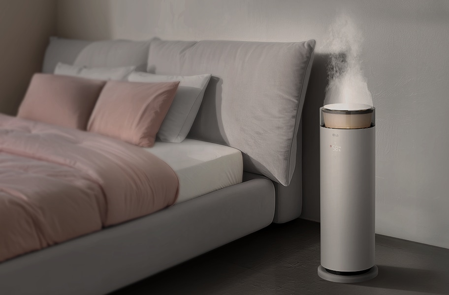 침실에 LG 퓨리케어 오브제컬렉션 하이드로에센셜 제품이 놓여 있으며, 포근 가습 모드가 켜져 있고 가습무가 나오고 있습니다. 디스플레이에는 58%라는 숫자가 표시되어 있습니다. 침대에는 여러 개의 베개와 이불이 정돈되어 있습니다.