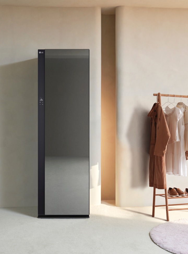 세련된 디자인의 LG 스타일러가 있으며, 옆에는 다양한 색상의 옷이 걸려 있는 옷걸이가 있습니다. 파란색 벨벳 소재의 의자가 놓여 있고, 벽에는 조명이 설치된 벽감이 있습니다. 바닥은 밝은 색상의 카펫으로 덮여 있습니다.