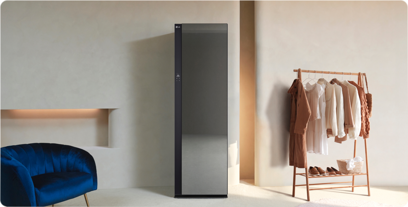 세련된 디자인의 LG 스타일러가 있으며, 옆에는 다양한 색상의 옷이 걸려 있는 옷걸이가 있습니다. 파란색 벨벳 소재의 의자가 놓여 있고, 벽에는 조명이 설치된 벽감이 있습니다. 바닥은 밝은 색상의 카펫으로 덮여 있습니다.