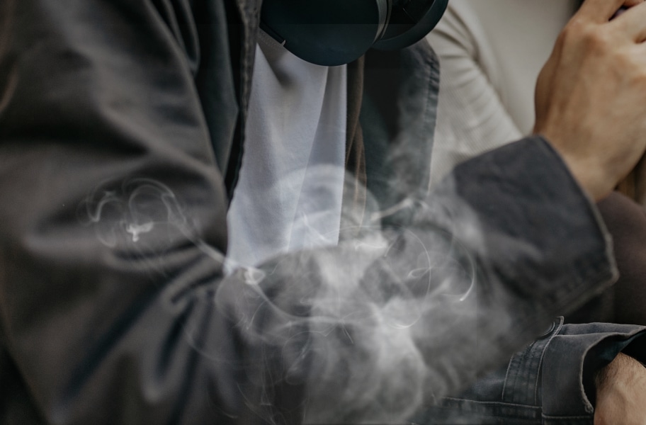 사람이 검은색 재킷을 입고 흰색 셔츠를 입고 있으며, 손에는 무언가를 쥐고 있습니다. 주변에는 연기가 피어오르고 있습니다.