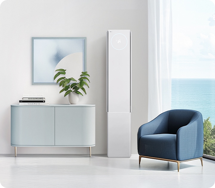 밝은 거실에 하얀색 2in1 에어컨이 설치되어 있으며, 에어컨 상단에는 24라는 숫자가 표시되어 있습니다. 에어컨 옆에는 푸른색의 안락의자가 놓여 있고, 그 옆에는 바다 풍경이 보이는 큰 창문이 있습니다. 왼쪽에는 식물이 놓인 작은 회색 수납장이 있으며, 그 위에는 두 권의 책과 함께 추상적인 그림이 걸려 있습니다.