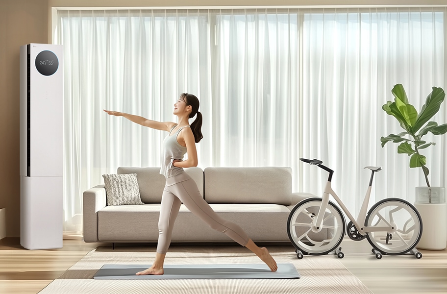 LG 휘센 AI 오브제컬렉션 뷰I 프로 에어컨이 왼쪽에 위치해 있으며, 에어컨의 디스플레이에는 24.5℃와 55%라는 숫자가 표시되어 있습니다. 에어컨 옆에는 밝은 색상의 소파가 놓여 있고, 소파 앞에는 요가 매트 위에서 운동하는 여성이 있습니다. 그녀는 오른팔을 앞으로 뻗고 왼쪽 다리를 뒤로 들어 올린 자세를 취하고 있습니다. 소파 오른쪽에는 실내 자전거가 놓여 있으며, 그 옆에는 화분에 심어진 녹색 식물이 보입니다. 배경으로는 커튼이 드리워진 큰 창문이 있습니다.
