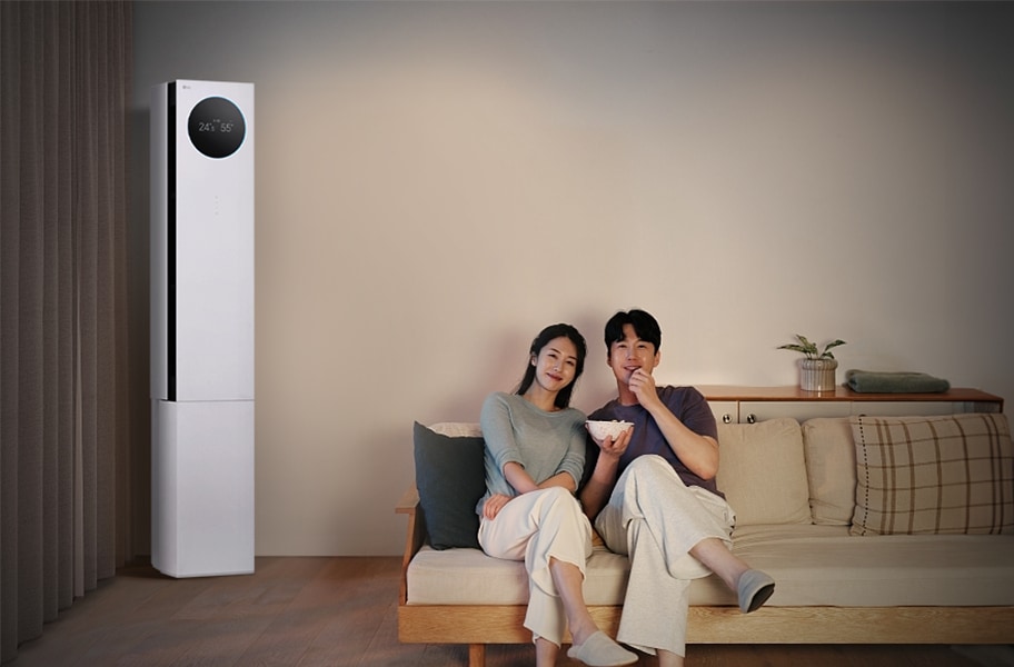 LG 휘센 AI 오브제컬렉션 뷰I 프로 에어컨이 왼쪽에 세워져 있으며, 원형 디스플레이에는 24.5℃와 55%라는 숫자가 표시되어 있습니다. 오른쪽에는 소파에 앉아 있는 두 사람이 보이며, 남성은 팝콘을 들고 있습니다. 소파 뒤쪽에는 수납장이 놓여 있고 그 위에는 식물과 수건이 놓여 있습니다.