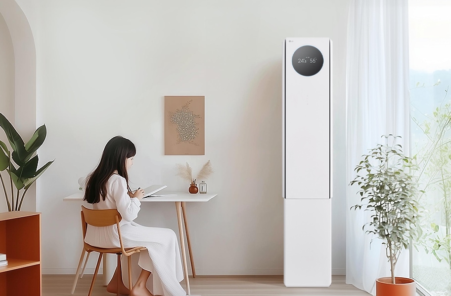 에어컨 상단에는 원형 디스플레이가 있으며, 24.5°와 55%라는 숫자가 표시되어 있습니다. 왼쪽에는 흰색 테이블과 나무 의자에 아이가 앉아 있으며, 테이블 위에는 책과 작은 장식품이 있습니다. 벽에는 식물 액자가 걸려 있으며, 오른쪽에는 커튼이 드리워진 창문과 화분이 보입니다.