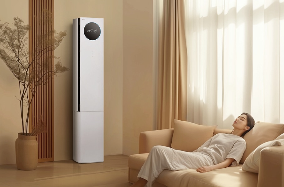 에어컨 상단에는 원형 디스플레이가 있으며, 24.5℃와 55%라는 숫자가 표시되어 있습니다. 에어컨 옆에는 나무 소재의 장식 패널과 화분이 놓여 있으며, 오른쪽에는 밝은 색상의 커튼이 드리워진 창문이 있습니다. 창문 앞에는 베이지색 소파가 놓여 있고, 소파에 한 여성이 편안하게 누워 있습니다. 실내는 따뜻한 색조로 꾸며져 있으며, 자연광이 부드럽게 들어오고 있습니다.