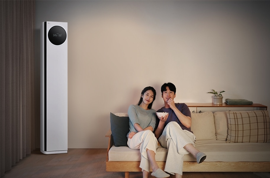 LG 휘센 AI 오브제컬렉션 타워I 에어컨이 왼쪽에 세워져 있으며, 원형 디스플레이에는 24.5℃와 55%라는 숫자가 표시되어 있습니다. 오른쪽에는 소파에 앉아 있는 두 사람이 보이며, 남성은 팝콘을 들고 있습니다. 소파 뒤쪽에는 수납장이 놓여 있고 그 위에는 식물과 수건이 놓여 있습니다.