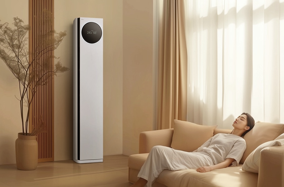 에어컨 상단에는 원형 디스플레이가 있으며, 24.5℃와 55%라는 숫자가 표시되어 있습니다. 에어컨 옆에는 나무 소재의 장식 패널과 화분이 놓여 있으며, 오른쪽에는 밝은 색상의 커튼이 드리워진 창문이 있습니다. 창문 앞에는 베이지색 소파가 놓여 있고, 소파에 한 여성이 편안하게 누워 있습니다. 실내는 따뜻한 색조로 꾸며져 있으며, 자연광이 부드럽게 들어오고 있습니다.