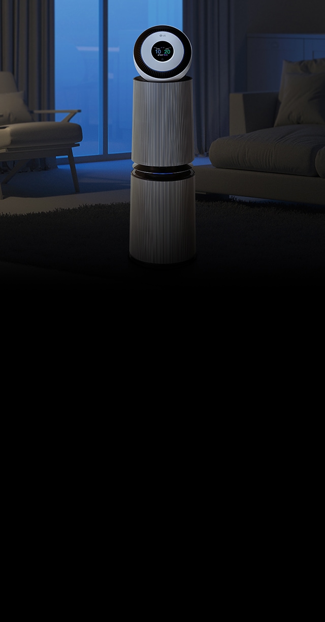 실내 거실에 공기청정기가 놓여 있습니다. 공기청정기는 원통형 디자인으로, 상단에 원형 디스플레이가 있으며 'LG' 로고가 보입니다. 주변에는 소파와 의자가 배치되어 있고, 창문을 통해 푸른빛이 들어옵니다.