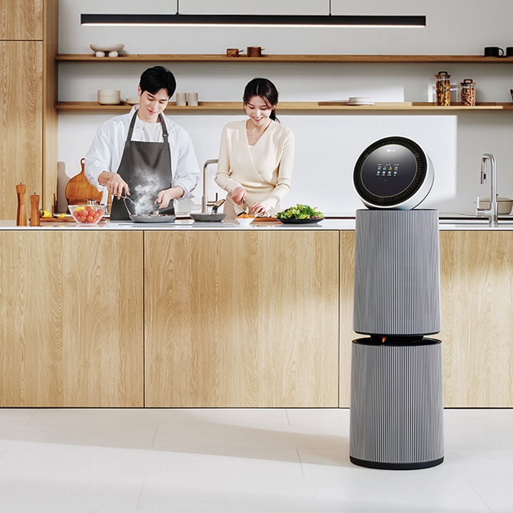 주방 공간에 LG 퓨리케어 AI 오브제컬렉션 360˚ 공기청정기가 있습니다. 나무 재질의 식탁과 의자가 놓여 있고 나무 장식의 주방 가구와 싱크대가 보입니다. 두 사람이 주방에서 요리를 준비하고 있는 모습이 함께 담겨 있습니다.