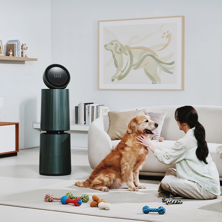 거실 공간에 LG 퓨리케어 AI 오브제컬렉션 360˚ 공기청정기가 놓여 있습니다. 한 여성이 앉아있는 강아지를 만지고 있으며 주변에는 강아지 장난감이 있습니다. 소파가 보이며, 벽에는 강아지 그림이 걸려 있고 선반에는 책과 장식품들이 놓여 있습니다.