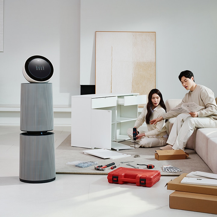 거실 공간에 LG 퓨리케어 AI 오브제컬렉션 360˚ 공기청정기가 놓여 있습니다. 가구를 조립하고 있는 두 사람의 모습이 보입니다. 주변에는 조립 중인 서랍장과 공구가 있습니다.