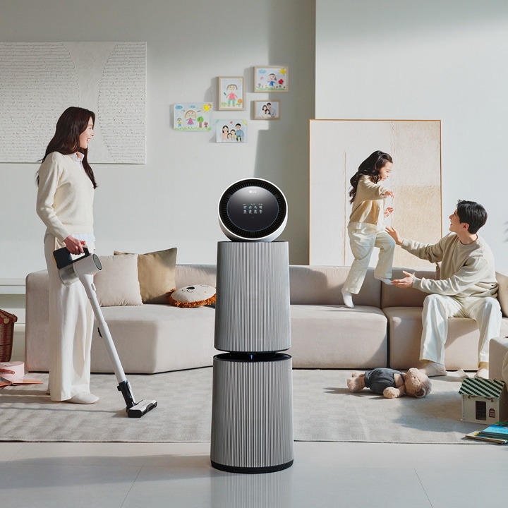 거실 공간에 LG 퓨리케어 AI 오브제컬렉션 360˚ 공기청정기가 있습니다. 소파에 앉아 있는 남성과 뛰어노는 아이, 그리고 청소기를 들고 있는 여성이 보입니다. 벽에는 그림이 걸려 있고, 바닥에는 화분과 장난감 등이 배치되어 있습니다.