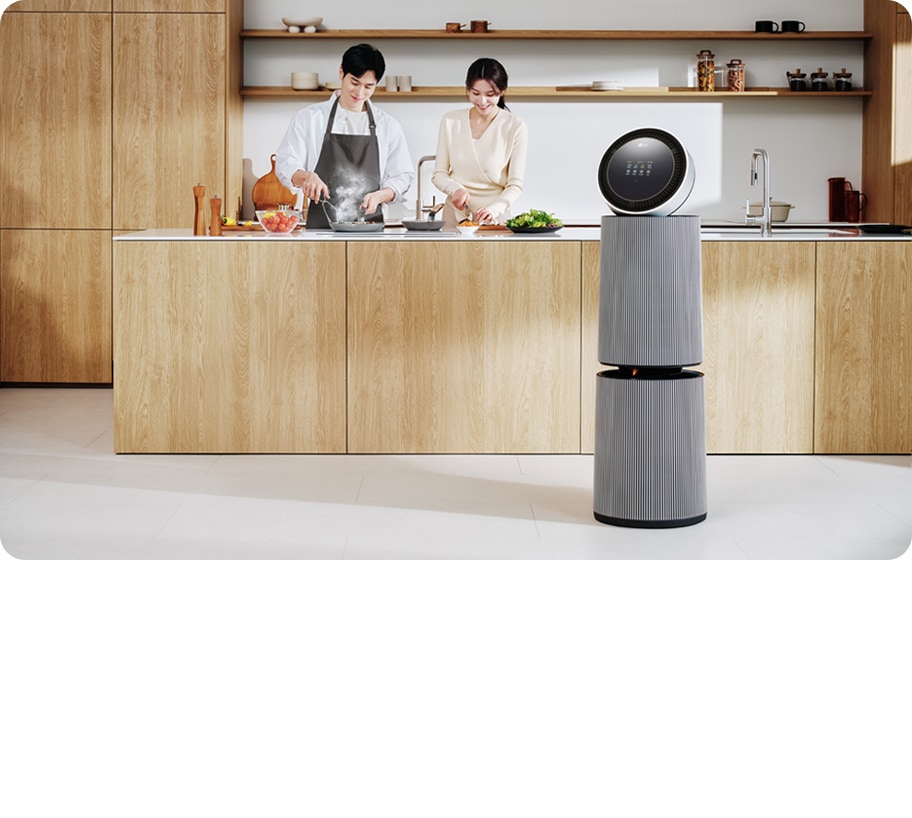 주방 공간에 LG 퓨리케어 AI 오브제컬렉션 360˚ 공기청정기가 있습니다. 나무 재질의 식탁과 의자가 놓여 있고 나무 장식의 주방 가구와 싱크대가 보입니다. 두 사람이 주방에서 요리를 준비하고 있는 모습이 함께 담겨 있습니다.