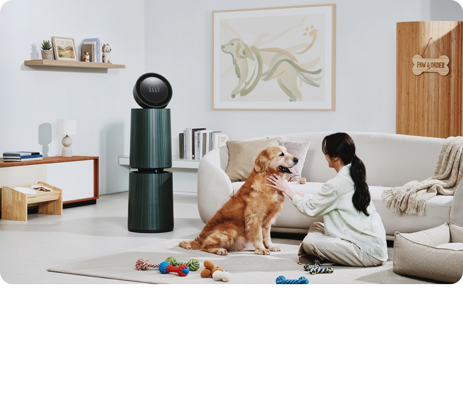 거실 공간에 LG 퓨리케어 AI 오브제컬렉션 360˚ 공기청정기가 놓여 있습니다. 한 여성이 앉아있는 강아지를 만지고 있으며 주변에는 강아지 장난감이 있습니다. 소파가 보이며, 벽에는 강아지 그림이 걸려 있고 선반에는 책과 장식품들이 놓여 있습니다.