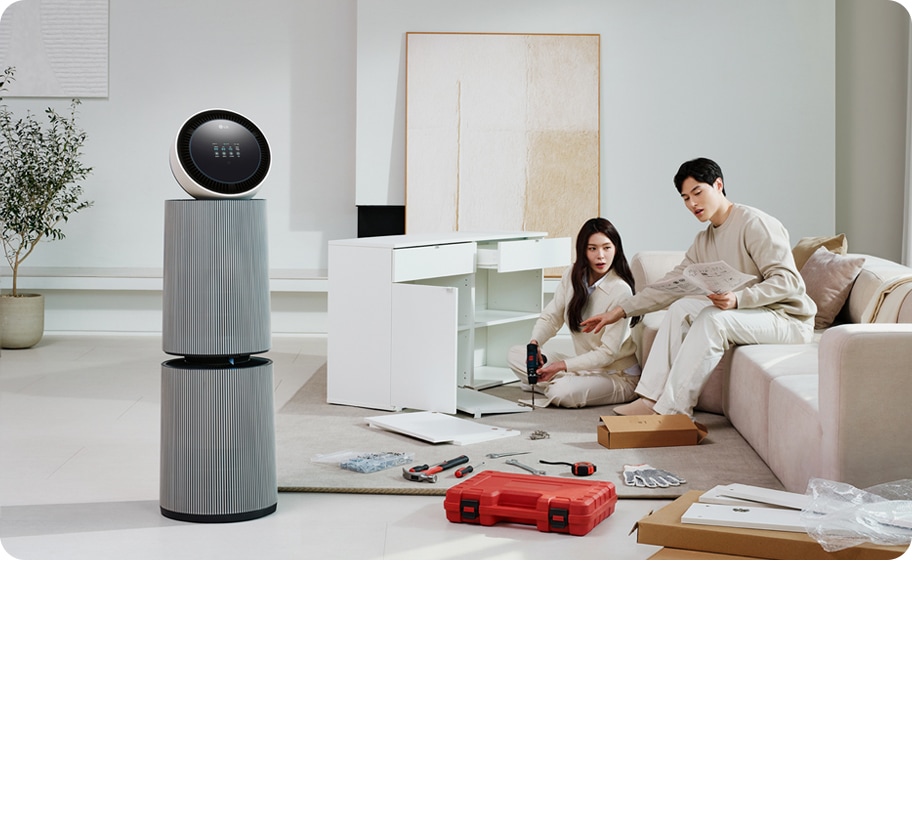 거실 공간에 LG 퓨리케어 AI 오브제컬렉션 360˚ 공기청정기가 놓여 있습니다. 가구를 조립하고 있는 두 사람의 모습이 보입니다. 주변에는 조립 중인 서랍장과 공구가 있습니다.