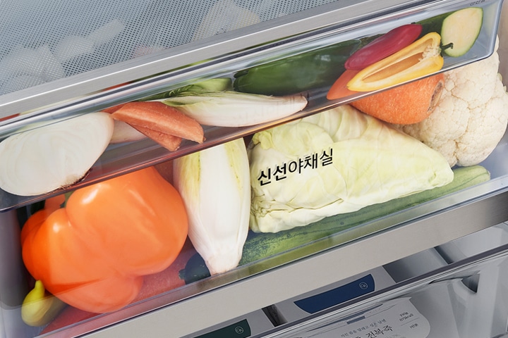 투명한 냉장고 서랍 안에 다양한 채소들이 담겨 있습니다. 양파, 당근, 양배추, 파프리카, 오이, 콜리플라워 등이 보이며, 서랍에는 신선야채실이라는 글자가 적혀 있습니다.