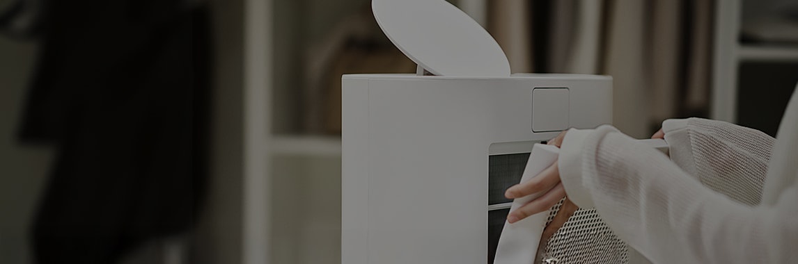 사람이 흰색 옷을 입고 제습기 필터를 교체하고 있는 모습입니다. 제습기는 흰색이며, 주변에는 옷이 걸려 있는 옷장이 보입니다.