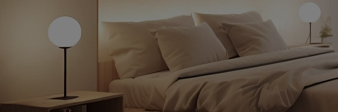 침대가 놓여 있으며, 베개와 이불이 깔끔하게 정돈되어 있습니다. 침대 양쪽에는 둥근 전구 모양의 스탠드 조명이 켜져 있고, 한쪽에는 작은 화분이 놓여 있습니다. 조명과 침구의 색상이 따뜻하고 아늑한 분위기를 연출하고 있습니다.