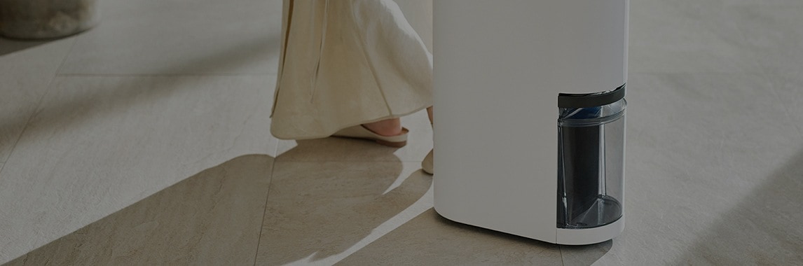 바닥에 긴 드레스를 입은 사람이 서 있고, 흰색 가전제품이 보입니다. 제품에는 투명한 부분이 있어 내부가 약간 보입니다. 바닥은 밝은 색상의 타일로 되어 있습니다.