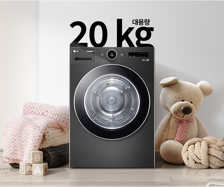 검정색 세탁기가 있으며, 상단에 20kg 대용량이라는 문구가 있습니다. 세탁기 옆에는 곰 인형과 두꺼운 니트 담요가 놓여 있고, 바닥에는 알파벳 블록이 있습니다. 