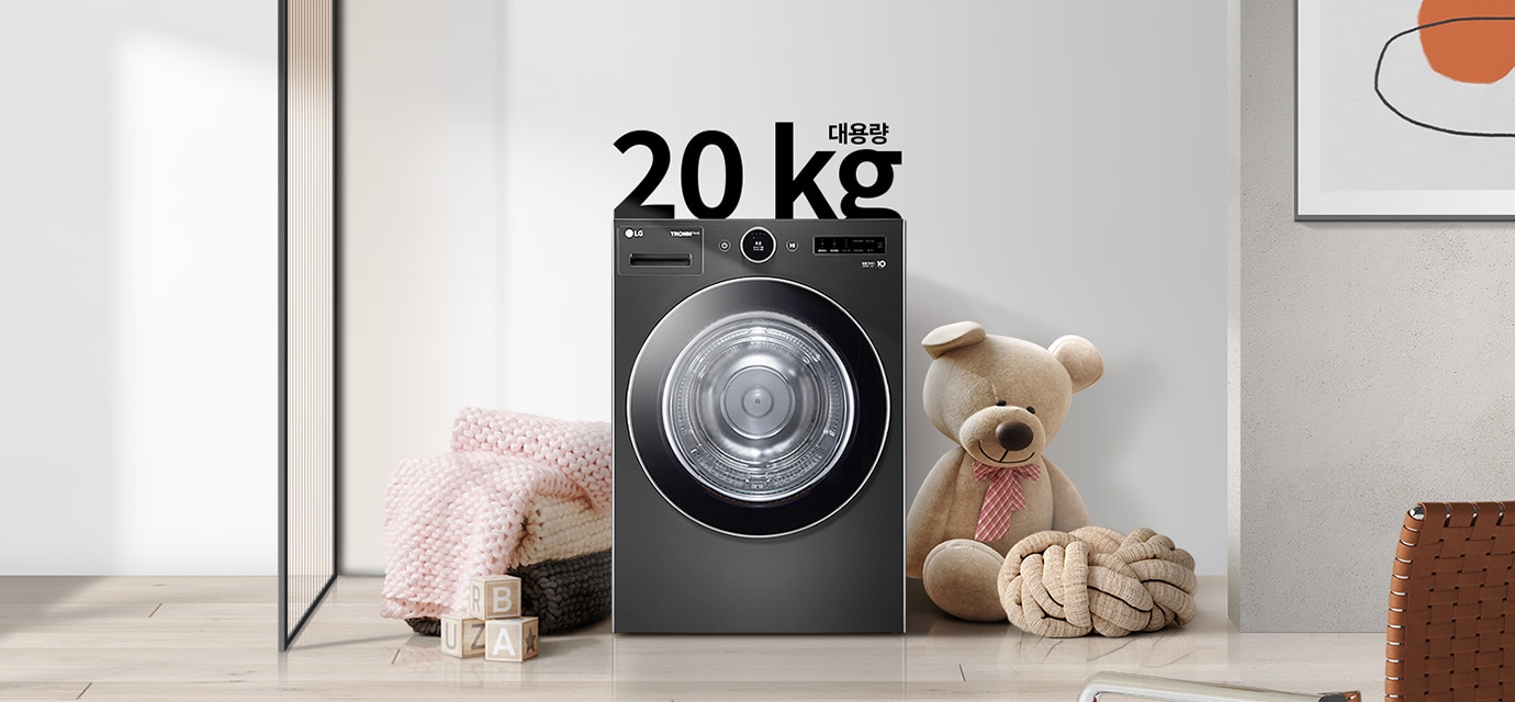 검정색 세탁기가 있으며, 상단에 20kg 대용량이라는 문구가 있습니다. 세탁기 옆에는 곰 인형과 두꺼운 니트 담요가 놓여 있고, 바닥에는 알파벳 블록이 있습니다. 
