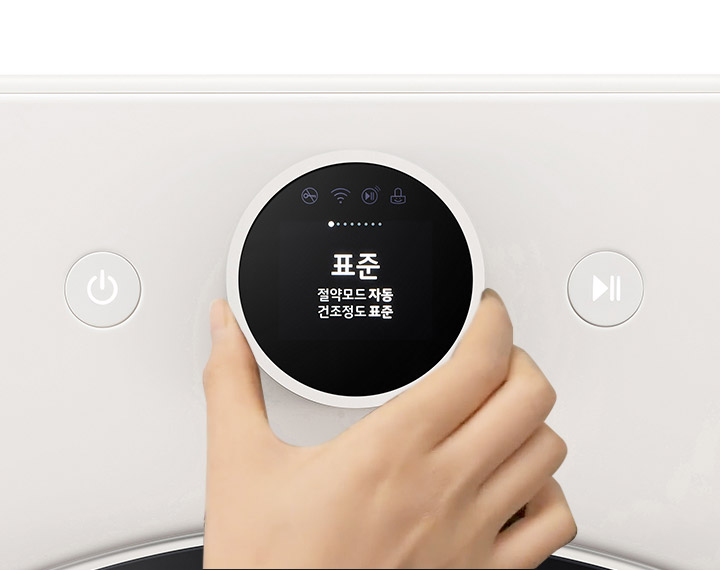 손이 조작하는 세탁기 조작부가 보입니다. Objet라는 로고가 있으며, 전원 버튼과 재생 버튼이 있습니다. 원형 디스플레이에는 표준, 절약모드 자동, 건조정도 표준이라는 텍스트가 표시되어 있습니다. 콘덴서케어, 절약모드, 건조정도, 건조시간, 다림질알림이라는 텍스트가 보입니다.