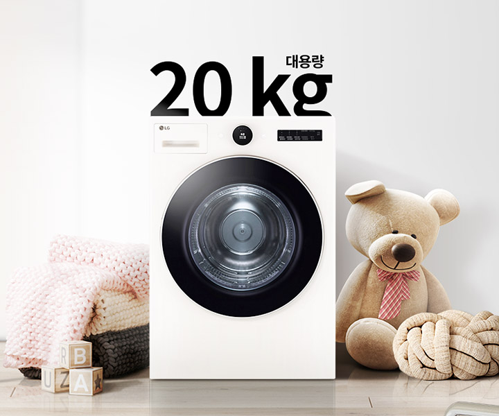세탁기가 놓여 있으며, 상단에 대용량 20kg이라는 텍스트가 있습니다. 세탁기 주변에는 분홍색 니트 담요가 담긴 바구니와 곰 인형, 장난감 블록이 있습니다. 벽에는 추상적인 그림이 걸려 있습니다. 바닥은 나무 마루로 되어 있습니다.